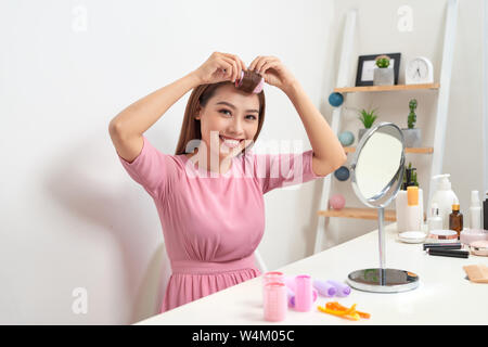 Head and shoulders portrait of beautiful Asian woman wearing hair curlers looking in mirror with wide smile, home interior on background Stock Photo