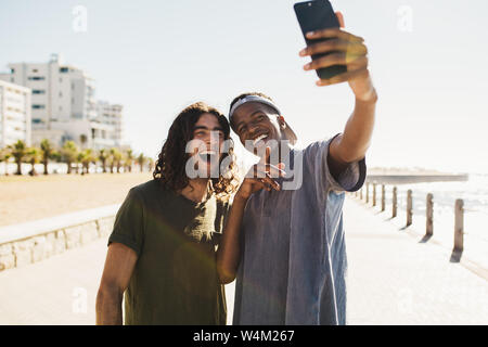 Two young mixed race men making a selfie by seaside promenade. Happy young friends taking a self portrait with smart phone outdoors. Stock Photo