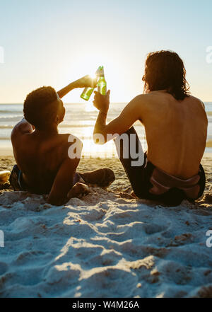Rear view of two young friends sitting on the beach and toasting beers during sunset. Young people partying in the beach with beers.