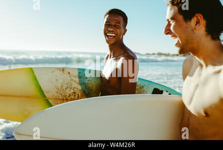 Happy young man with a friend walking on the beach carrying surfboards. Cheerful young friends on vacation at the sea.