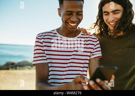 Two young friends outdoors looking at mobile phone and smiling. Young men using a smart phone by the beach. Stock Photo