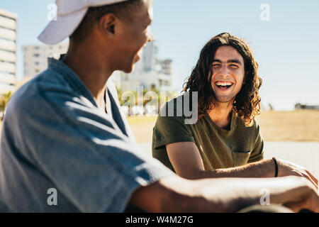 Young man smiling while talking with his friend outdoors. Two young men sitting outdoors and smiling at each other. Stock Photo