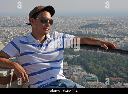 Handsome young man wearing a baseball cap and sun glasses sitting in the sun with the city of Athens in the background Stock Photo