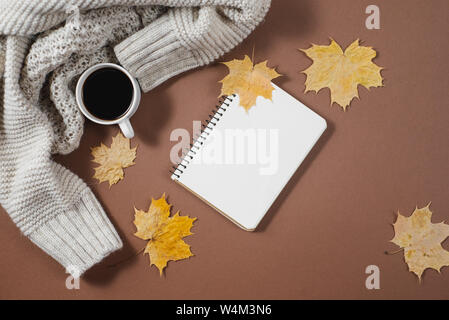 Workspace with golden maple leaves, notebook, coffee cup, sweater on brown background. Creative composition. Autumn or Winter concept. Flat lay, top v Stock Photo