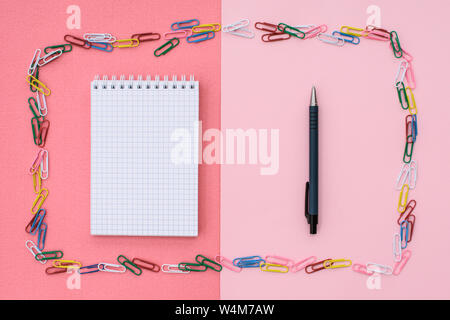 Notebook in cage with a black pen in a frame of colored paper clips on a double background - pink and coral. Concept back to school. Stock Photo