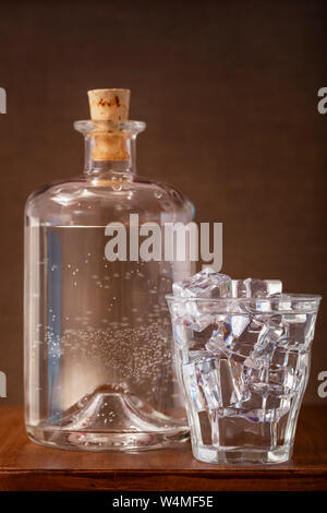 Nostalgia still of a glass of ice cold water with ice cubes and a vintage bottle with cork on  a wooden table Stock Photo