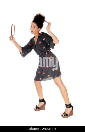 Lovely young woman standing in a black dress with flower print holding a mirror in her hand fixing her black curly hair, looking into the camera, isol Stock Photo