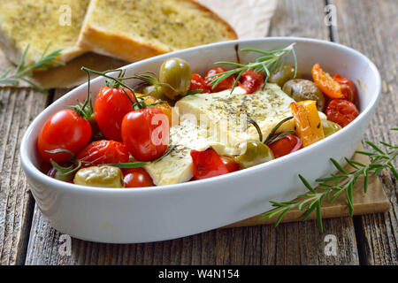 Warm Greek appetizer: Baked feta cheese with green olives, cherry tomatoes,  peppers, onions  and olive oil served with roasted herb baguette Stock Photo