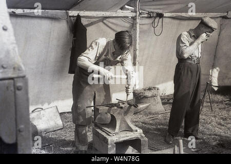 1950s, historical, a farrier working on a horseshoe using a hammer and an anvil in a tent outside at a country fair, England, UK. Traditionally the jobs of a farrier and a blacksmith were the same, with the word 'farrier' deriving from the french word for blacksmith (ferrier). Stock Photo