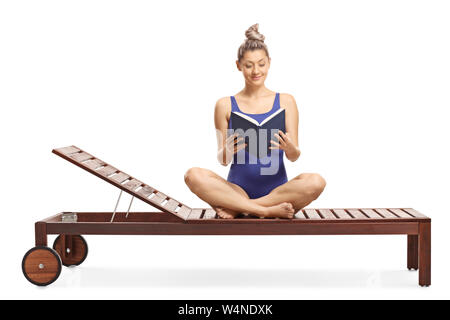 Young female in a swimming suit sitting on a sunbed and reading a book isolated on white background Stock Photo