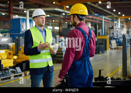 Two engineers in working uniforms and protective helmets have a work related conersation involving blueprints brought along. Stock Photo