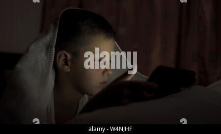 A happy boy lies in bed under a blanket and plays on a tablet in a game in the dark. The face of the child is lit by a bright monitor Stock Photo