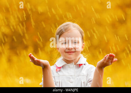 Portrait of a cute charming little girl standing in autumn park in the rain. The child stretched out his hands and collects the drops in his palm. Blu Stock Photo