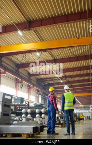 A view of two engineers in the uniform and helmets among the industrial equipment of the plant they are working in both talking to each other. Stock Photo