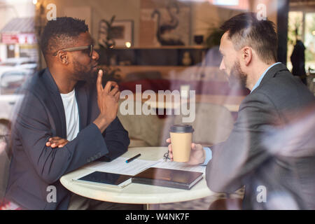 Two men in suits sit in the cafe, drink coffee and have business related conversations while looking through the documents. Stock Photo