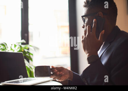 Afro-american in a suit working as a freelancer in the cafe with his laptop. And drinking tea at the same time as having a conversation on the phone. Stock Photo