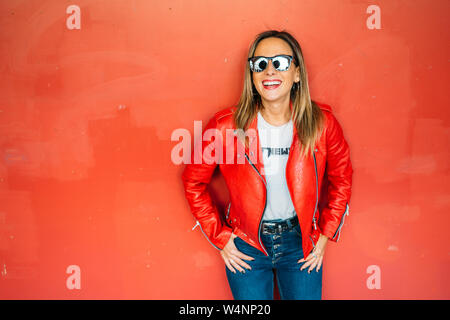 Attractive woman looking at camera and laughing with sunglasses and red leather jacket over red background. Rocker Girl in urban background. Stock Photo