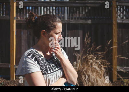 woman stands in front of fence in sunlight with thoughtful expression Stock Photo