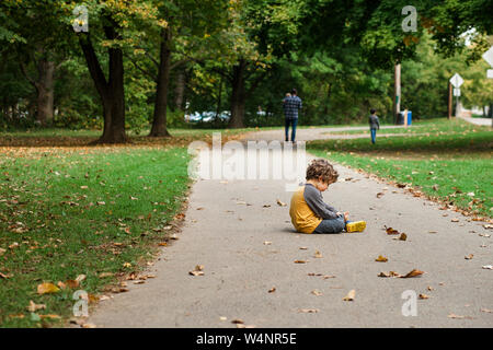 A small boy sits on a leaf-strewn path as his family walks on Stock Photo