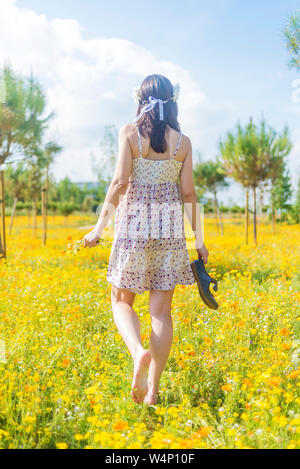 Rear view of a beautiful woman in dress walking through a field of flowers carrying her shoes