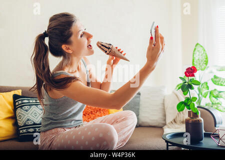 Happy young woman eating chocolate ice-cream in cone sitting on couch at home and taking selfie using smartphone. Stock Photo
