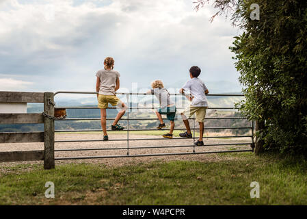 Three kids climbing on a gate with mountains in the distance Stock Photo