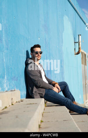 Portrait of stylish man sitting on stairs leaning on a blue wall while looking camera Stock Photo