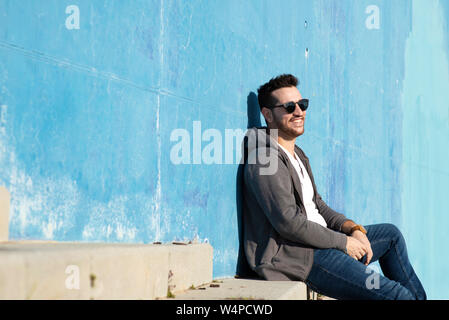 Portrait of stylish man sitting on stairs leaning on a blue wall while looking away and smiling Stock Photo