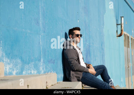 Portrait of stylish man sitting on stairs leaning on a blue wall while looking away Stock Photo