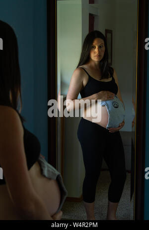 Pregnant woman late in the third trimester holding up baby clothing to her baby bump, looking anxious about the upcoming life change. Stock Photo