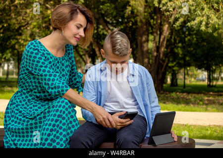 Mom and son using tablet and smartphone while sitting on nature. The family plays computer or searches the Internet, electronic devices and children Stock Photo