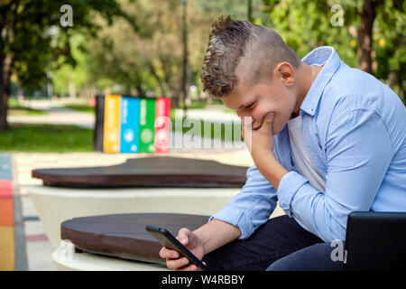 Problems of modern children. Young guy in the park with the phone upset. Depression is a mental disorder due to social networks. Online life Stock Photo