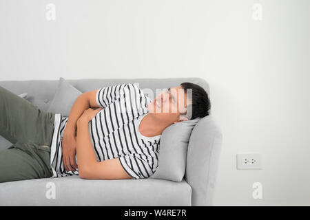 Terrible stomachache. Frustrated handsome young man hugging his belly and keeping eyes closed while lying on the couch at home Stock Photo