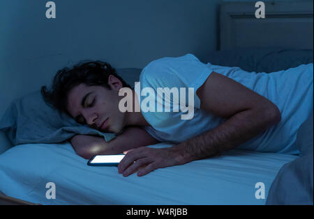 Addicted to social media young man falling asleep with smart mobile phone at night in bed. lifestyle portrait of man sleeping in dark bedroom with mob Stock Photo