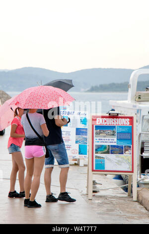 Vodice, Croatia - July 13, 2019: Family holding umbrellas in front of boat excursion billboards on a rainy summer day by the sea Stock Photo