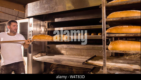 bakery worker taking out freshly baked breads with shovel from the professional oven at the manufacturing Stock Photo