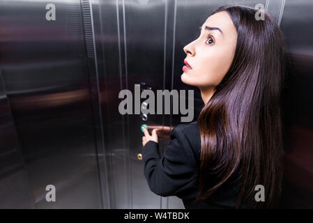 scared businesswoman, suffering from claustrophobia, looking up while pushing button in elevator Stock Photo