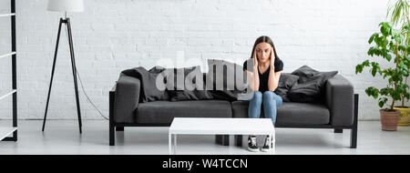 depressed young woman sitting on sofa in spacious room and holding hands near head Stock Photo