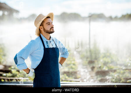 Portrait of a handsome farmer in blue workwear and straw hat standing on the farmland with automatic watering on the sunset Stock Photo