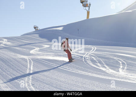 Snowboarder in red descends on snowy ski slope prepared by grooming machine at sunny winter day Stock Photo