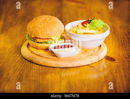 Burger with cheese meat and salad. Cheat meal. Delicious burger with sesame seeds. Fast food concept. Burger menu. High calorie snack. Hamburger and french fries and tomato sauce on wooden board. Stock Photo