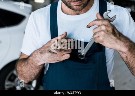 bearded auto mechanic with mud on hands holding hand wrench in car repair station Stock Photo