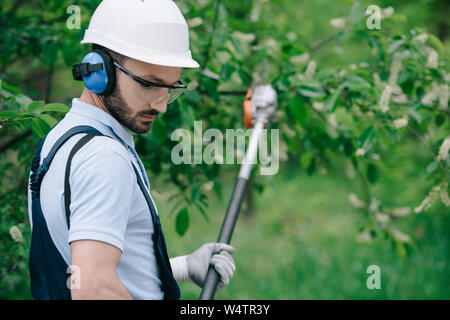 selective focus of pensive gardener in helmet and hearing protectors trimming trees with telescopic pole saw in park Stock Photo