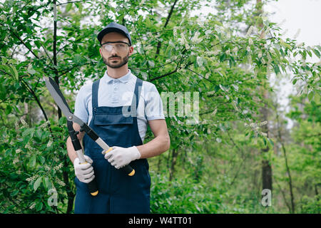 serious gardener in overalls, cap and protective glasses holding trimmer and smiling at camera Stock Photo