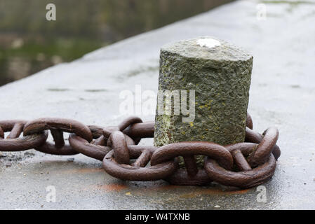 Large rusty old fishing boat chain looped around a concrete mooring on a wet harbour (harbor) wall. Stock Photo