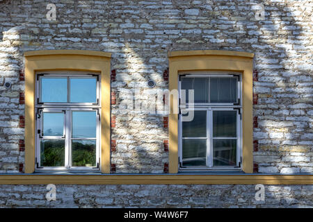 Two rectangular windows with yellow wooden frames on the background of a stone wall with a shadow from the tree. From the window series of the world. Stock Photo