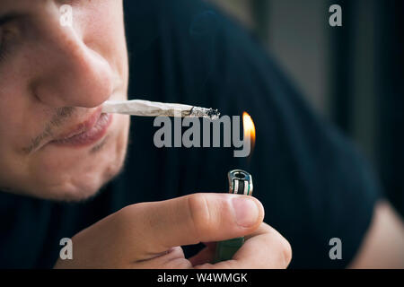 A young guy in a blue t-shirt with a bristle on his face, holding a cigarette in his mouth with his lips, lights it with fire from a gas pocket green Stock Photo