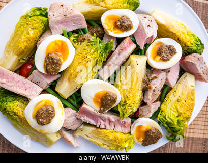 Version of salad Nicoise on white plate.  Hard boiled eggs and olive dressing, baby gem lettuce, boiled potatoes & tuna steak slices