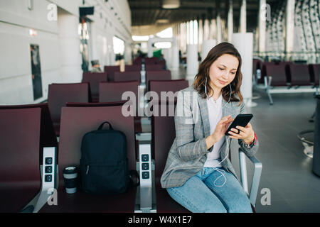 Young girl at the airport waiting for her departure. Scrolling smartphone in headphones. A cup of coffee and backpack beside. Stock Photo