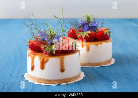 Two round no bake strawberry mini cheesecakes with salt caramel and halfs of strawberries, embellished with real blue blossoms. Blue wooden table. Stock Photo